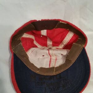 May include: Red and blue baseball cap with a white interior. The cap has a brown leather band with a white stitched line. The inside of the cap has black writing that says "M. Howard, Evergreen 1288".