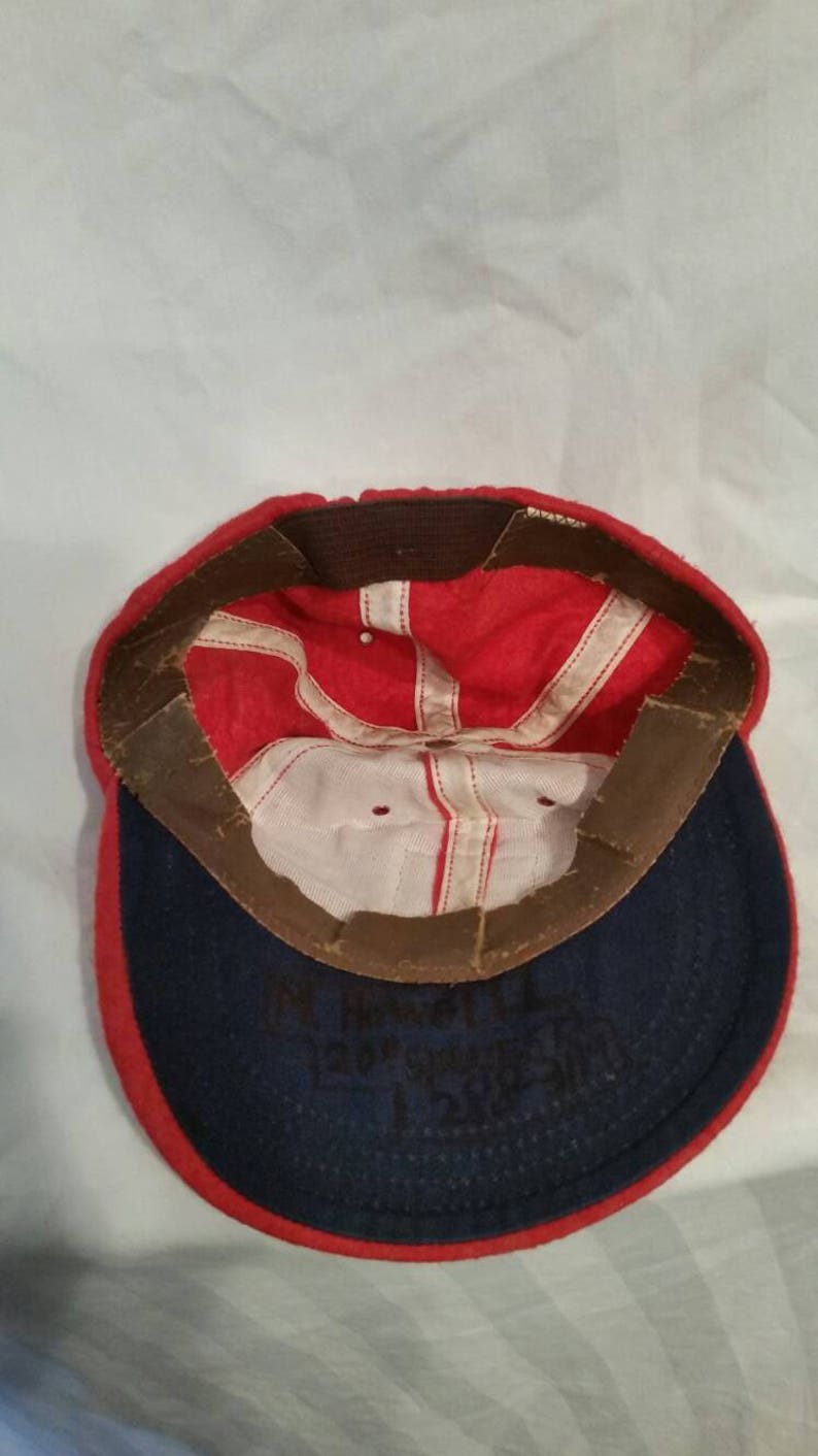 May include: Red and blue baseball cap with a white interior. The cap has a brown leather band with a white stitched line. The inside of the cap has black writing that says "M. Howard, Evergreen 1288".