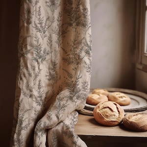 Può includere: Una tenda beige con un motivo ripetuto di illustrazioni botaniche grigie è appesa accanto a un tavolo di legno. Diverse pagnotte di pane rotonde sono sul tavolo e un vassoio di legno. La tenda è drappeggiata, rivelando la trama del tessuto.