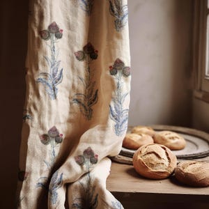 Op de afbeelding: Een beige linnen gordijn met een herhalend patroon van paarse en blauwe distelbloemen. Het gordijn is gedrapeerd over een houten oppervlak, met verschillende ronde broden.