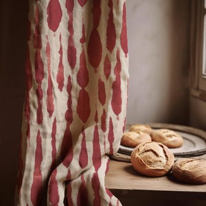 Può includere: Una tenda di lino beige con un motivo ripetuto di pennellate rosse verticali. Diverse pagnotte di pane rotonde e croccanti sono su un tavolo di legno e un vassoio rotondo di legno.