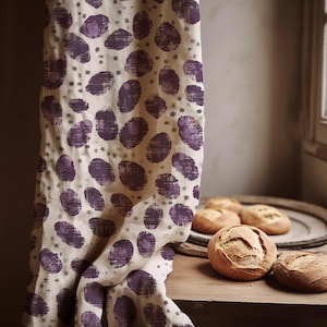 Può includere: Un tessuto beige con un motivo ripetuto di grandi ovali viola strutturati e piccoli punti grigi. Il tessuto è drappeggiato su una superficie di legno, con diversi panini rotondi e croccanti.