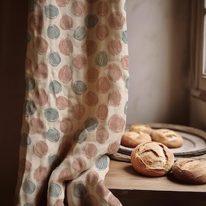 Può includere: Tessuto beige con un motivo ripetuto di forme ovali rosa e blu. Il tessuto è drappeggiato su una superficie di legno, accanto a un piatto con panini freschi. La scena è illuminata dalla luce naturale.
