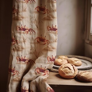 May include: A beige fabric curtain featuring a repeating pattern of reddish-brown crabs and tall grass stalks. The textile has a textured weave and hangs vertically next to a wooden surface holding several round, crusty bread rolls.