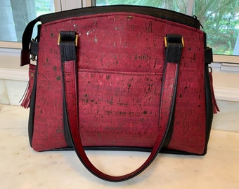 May include: A burgundy and black tote bag with a cork-like texture. The bag features black handles, a black shoulder strap, and gold-colored hardware. A small pocket is visible on the front. The bag has a zipper closure and a tassel.