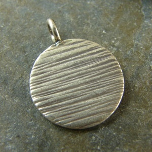 May include: A silver-colored pendant with a circular design. The pendant has a textured surface with horizontal lines. It features a small loop at the top for attaching to a chain. The pendant is likely made of metal.