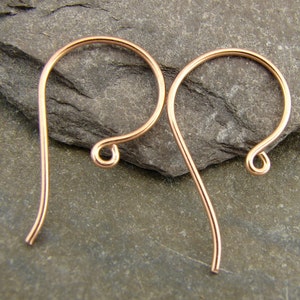 May include: Two copper-colored earring hooks are displayed against a gray stone surface. Each hook has a curved top and a looped bottom, designed to be inserted into pierced ears. The hooks are made of thin, smooth metal.