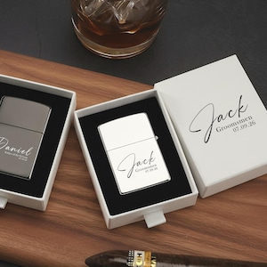 May include: Two personalized lighters in white boxes with names and dates, displayed on a wooden board. One lighter is silver, the other is white. Cigars and a glass of liquid are also present.