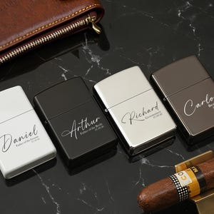 May include: Four personalized lighters in white, black, silver, and brown, each engraved with a name and date. A brown leather wallet and a cigar with a gold holder are also visible on a black marble surface.