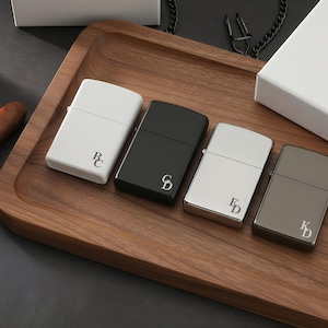 May include: Four personalized lighters in white, black, silver, and gunmetal gray, each with initials. The lighters are arranged on a wooden tray with a cigar and a white box in the background. A chain is also visible.
