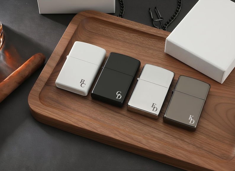 May include: Four personalized lighters in white, black, silver, and gunmetal gray, each with initials. The lighters are arranged on a wooden tray with a cigar and a white box in the background. A chain is also visible.