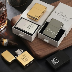 May include: An assortment of personalized lighters in gold, silver, and black, some in white boxes. The gold lighter is open, revealing the interior. The lighters are engraved with initials, names, and a mountain design. A cigar and a glass of whiskey are in the background.