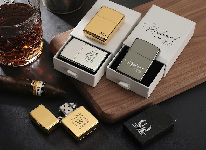 May include: An assortment of personalized lighters in gold, silver, and black, some in white boxes. The gold lighter is open, revealing the interior. The lighters are engraved with initials, names, and a mountain design. A cigar and a glass of whiskey are in the background.