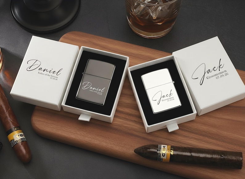 May include: Two personalized lighters in white boxes with names and dates, displayed on a wooden board. One lighter is silver, the other is white. Cigars and a glass of liquid are also present.