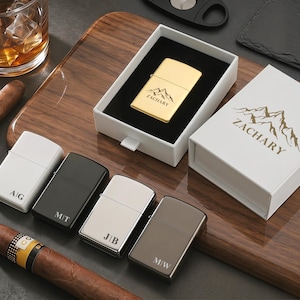 May include: An assortment of personalized lighters in various colors, including gold, black, white, and silver. One gold lighter is displayed in a white box with the name "ZACHARY" and a mountain graphic. Other lighters have initials engraved.