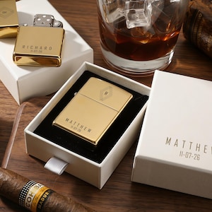 May include: Gold-colored lighters with personalized text and geometric designs are displayed in white boxes. One lighter is open, revealing the flint and wick. A glass of whiskey with ice and a cigar are also visible.