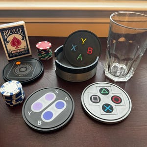 May include: A set of six black and white coasters with different video game controller button designs. The coasters are made of a hard, durable material and feature a variety of colors, including red, blue, green, and yellow. The coasters are perfect for protecting your furniture from spills and scratches.