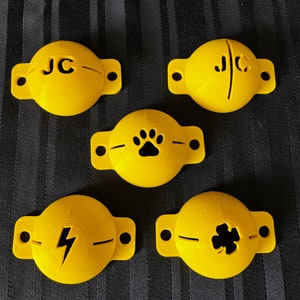 May include: Five yellow, circular objects with a flat, rectangular extension on each side. Each object has a different design: initials "JC", a paw print, a lightning bolt, and a four-leaf clover. The objects are likely decorative or functional.