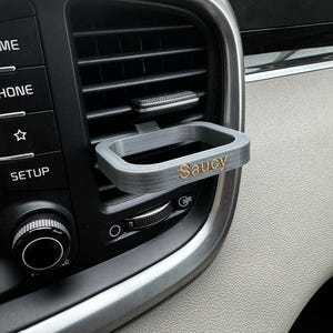 May include: A gray plastic car air vent holder with the word "Saucy" printed in gold lettering.