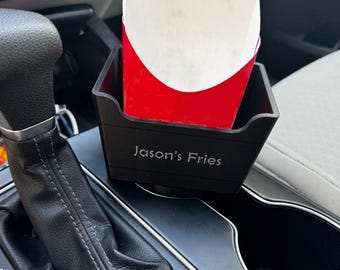 May include: A black car console french fry holder with the text "Jason's Fries" in a white font. The holder contains a red and white paper container filled with french fries. The holder is placed in a car's center console.