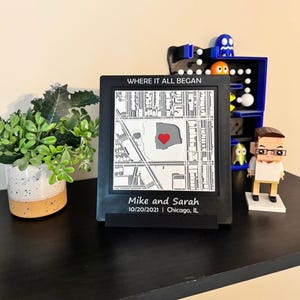 May include: A black framed map with the text "WHERE IT ALL BEGAN" and the names "Mike and Sarah" with a date and location. The map features a red heart marking a location. A small plant and figurines are also visible.