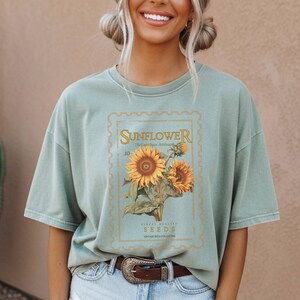 May include: Sage green t-shirt featuring a vintage-style sunflower graphic. The design includes the word "Sunflower" in a decorative font, along with the scientific name and the word "Seeds". The graphic is framed within a light blue border.