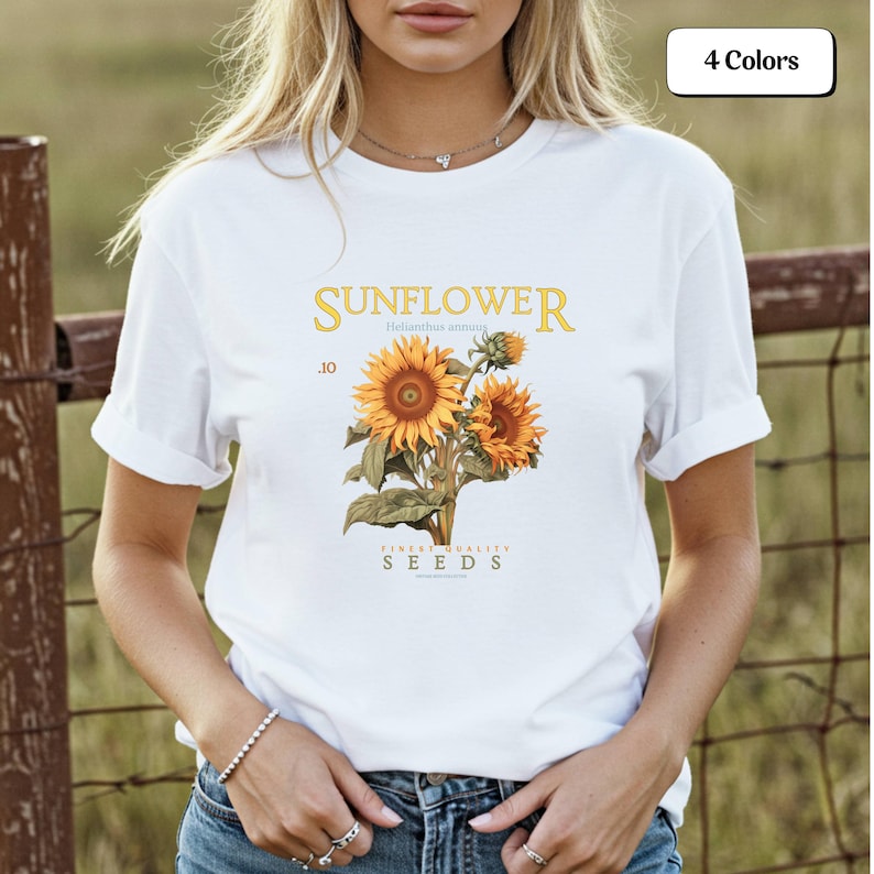 May include: White t-shirt featuring a sunflower design with the word "SUNFLOWER" in gold lettering. The graphic includes botanical illustrations of sunflowers and the text "FINEST QUALITY SEEDS".