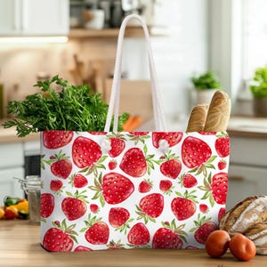 May include: White tote bag with a repeating pattern of red strawberries and green leaves. The bag has white rope handles and is filled with groceries, including fresh herbs and bread.