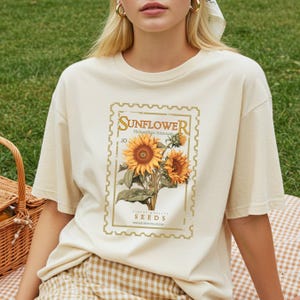 May include: A cream-colored short-sleeve t-shirt featuring a graphic print of a postage stamp design. The print displays two sunflowers with green leaves and the text "SUNFLOWER Helianthus Annuus .10 FINEST QUALITY SEEDS VINTAGE SEED-COLLECTIVE".
