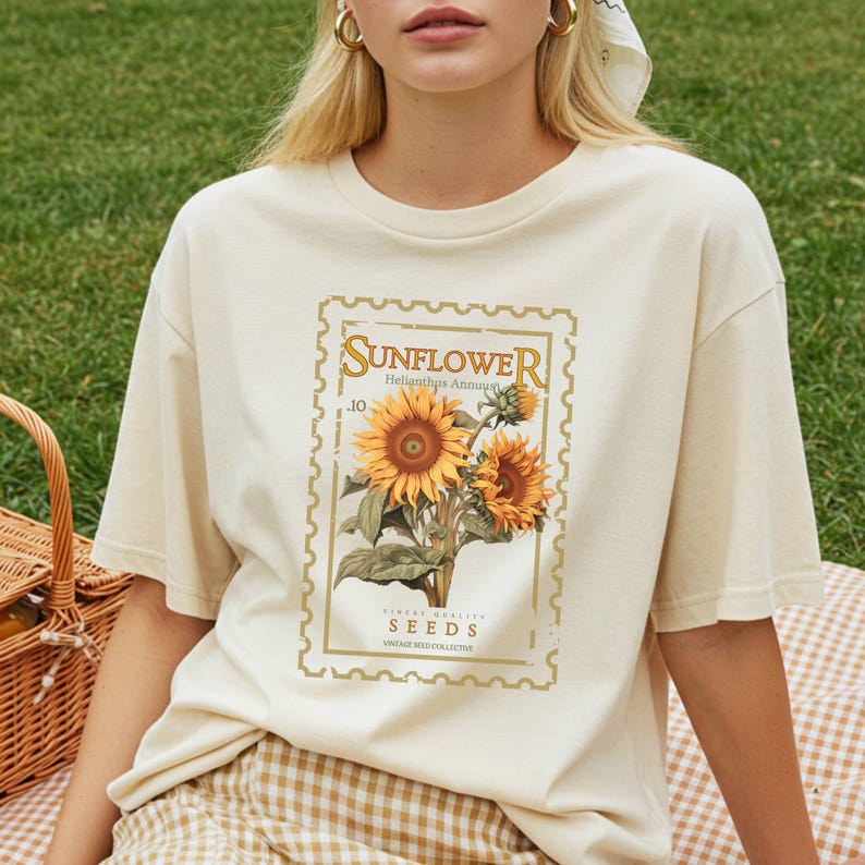 May include: A cream-colored short-sleeve t-shirt featuring a graphic print of a postage stamp design. The print displays two sunflowers with green leaves and the text "SUNFLOWER Helianthus Annuus .10 FINEST QUALITY SEEDS VINTAGE SEED-COLLECTIVE".