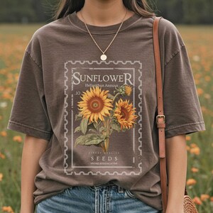 May include: Brown t-shirt featuring a vintage-style sunflower design within a postage stamp frame. The design includes the word "SUNFLOWER", the Latin name "Helianthus Annuus", and the word "SEEDS". The shirt has short sleeves and a crew neck.