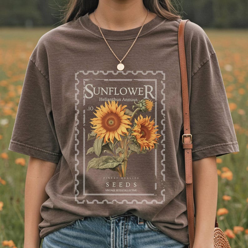 May include: Brown t-shirt featuring a vintage-style sunflower design within a postage stamp frame. The design includes the word "SUNFLOWER", the Latin name "Helianthus Annuus", and the word "SEEDS". The shirt has short sleeves and a crew neck.