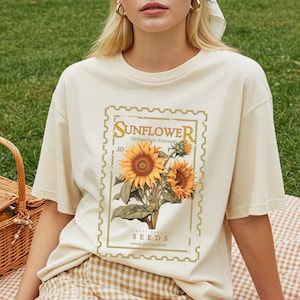 May include: Cream-colored t-shirt featuring a vintage-style sunflower design. The graphic includes the word "SUNFLOWER" in a decorative font, along with botanical details and the text "Finest Quality Seeds". The shirt has short sleeves and a relaxed fit.