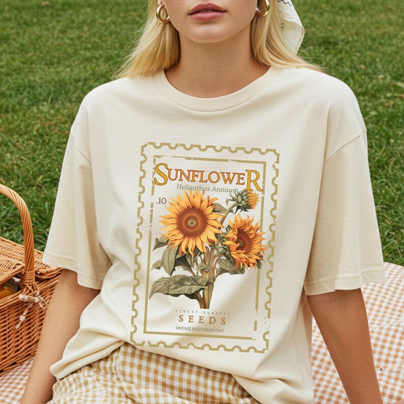 May include: Cream-colored t-shirt featuring a vintage-style sunflower design. The graphic includes the word "SUNFLOWER" in a decorative font, along with botanical details and the text "Finest Quality Seeds". The shirt has short sleeves and a relaxed fit.