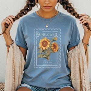 May include: A blue short-sleeved t-shirt featuring a graphic print of a vintage postage stamp design. The graphic displays two yellow sunflowers with green leaves and the text Sunflower, Helianthus Annuus, 10, Seeds, Vintage Seed Collective.