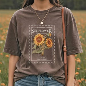 May include: A brown short-sleeve t-shirt featuring a graphic print of a postage stamp design with two yellow sunflowers. The text reads SUNFLOWER Helianthus Annuus .10 SEEDS VINTAGE SEED COLLECTIVE. A thin gold chain necklace is worn over the shirt.