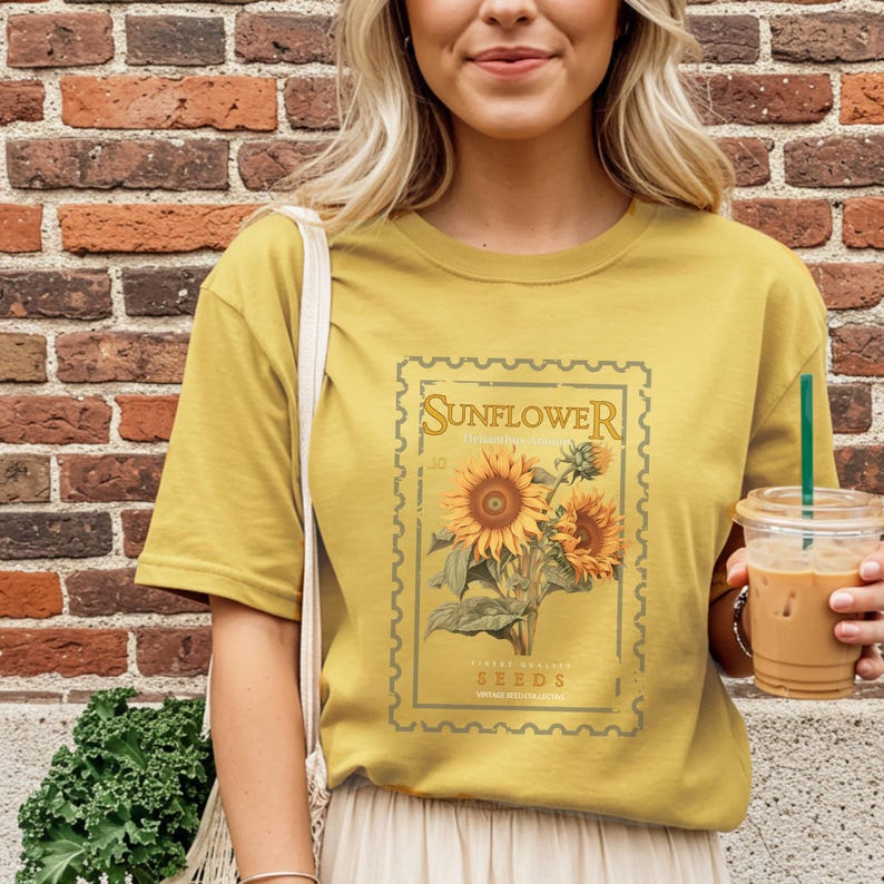 May include: A mustard yellow short-sleeve t-shirt featuring a rectangular graphic designed like a postage stamp. The graphic displays a detailed illustration of sunflowers with the text Sunflower and Seeds printed in a vintage-style font.