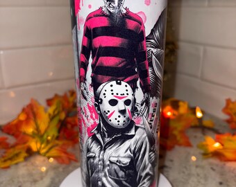 May include: A stainless steel tumbler featuring a horror movie theme. The design includes illustrations of Freddy Krueger in a striped sweater and Jason Voorhees in a hockey mask, with pink accents and a white background.