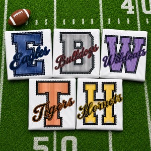 May include: Five white t-shirts featuring large, vertically striped block letters with embroidered edges. The letters E, B, W, T, and H are paired with the cursive team names Eagles, Bulldogs, Wildcats, Tigers, and Hornets in matching colors.