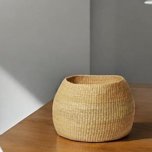 May include: A natural woven basket with a round, slightly oval shape. The basket is made of a light brown material and has a smooth, textured surface.