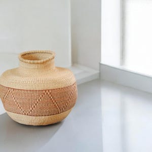 May include: A large, round, woven basket with a natural beige color and a brown geometric pattern around the middle.