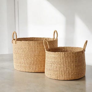 May include: Two natural woven storage baskets with handles. The larger basket is in the background and the smaller basket is in the foreground.