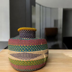 May include: A woven storage basket with a colorful geometric pattern in shades of pink, green, blue, and yellow. The basket has a yellow rim and a dark purple base.