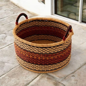 May include: A round woven basket with two handles. The basket features horizontal stripes in shades of tan, black, and reddish-brown. The handles are dark with red accents. The basket is sitting on a stone surface.