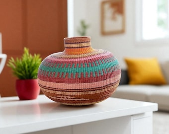 May include: A handwoven, colorful basket vase with a round body and a narrow neck. The vase features horizontal bands of brown, orange, pink, and teal. The vase is sitting on a white surface.