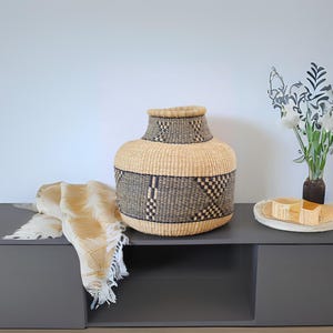 May include: A large, woven basket with a black and tan geometric pattern sits on a dark gray shelf. The basket is made of natural materials and has a round, wide base.