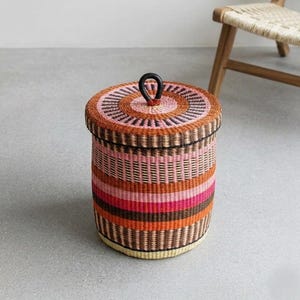 Könnte beinhalten: Ein runder, gewebter Aufbewahrungskorb mit Deckel. Der Korb hat eine Farbpalette aus rosa, orangefarbenen und braunen Streifen. Der Deckel hat ein Spiraldesign und einen schwarzen Griff. Der Korb ist etwa 30 cm hoch.
