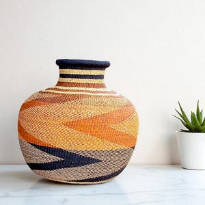 May include: A woven vase with a rounded body and a narrow neck. The vase features a geometric pattern in shades of orange, brown, and black. The vase is sitting on a white surface next to a small potted plant.