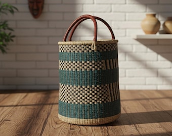 May include: A cylindrical woven basket with a teal and black checkered pattern and brown leather handles. The basket is made of natural fibers and sits on a wooden floor. The background includes a white brick wall and decorative items.
