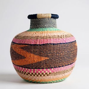 May include: A handwoven basket with a wide, rounded body and a narrow neck. The basket features a vibrant design with bands of pink, orange, green, and black. The top rim is dark blue, and the base has a checkered pattern.